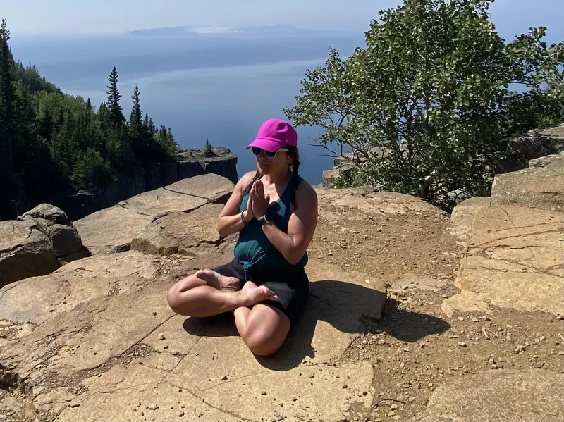 Lori Schmidt in seated meditation on a cliffside hiking trail, trees and skyline behind